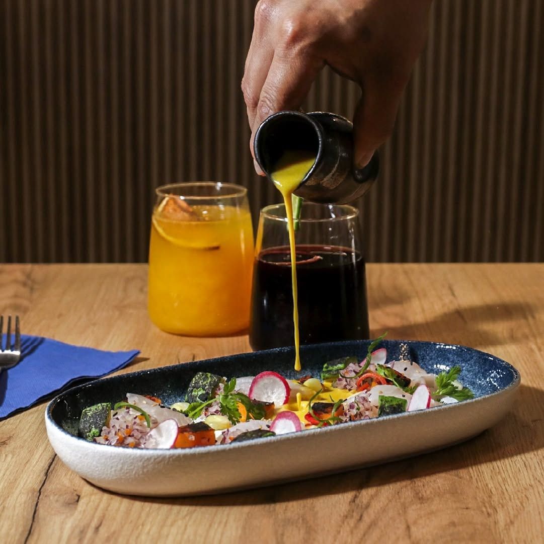 Una mano vierte el aliño sobre una ensalada colorida en una mesa de madera, con dos vasos y un frasco al fondo