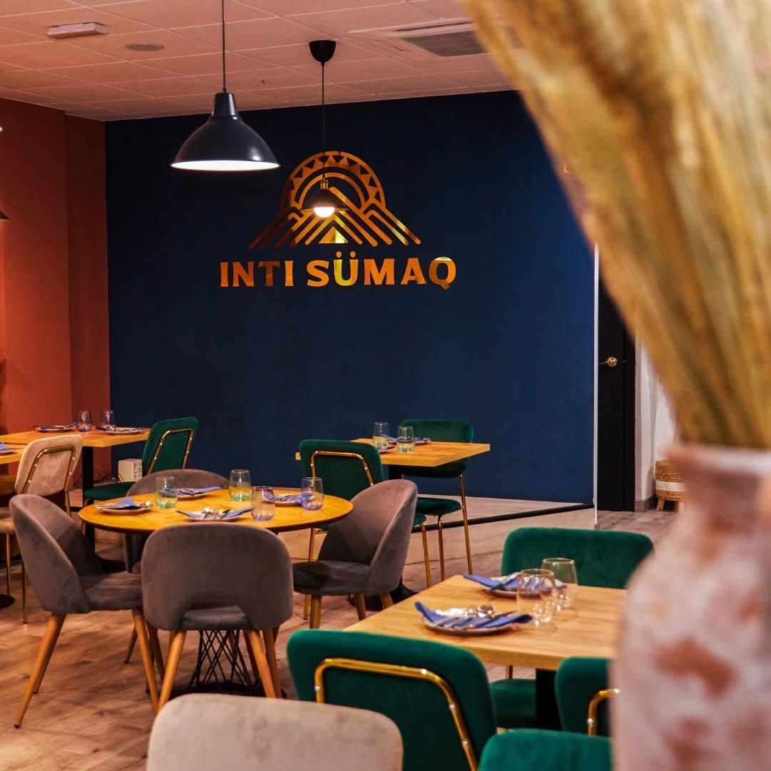 Interior de un restaurante moderno con sillas turquesa y doradas, mesas preparadas y un letrero que dice “INTI SUMAO” en una pared azul oscuro