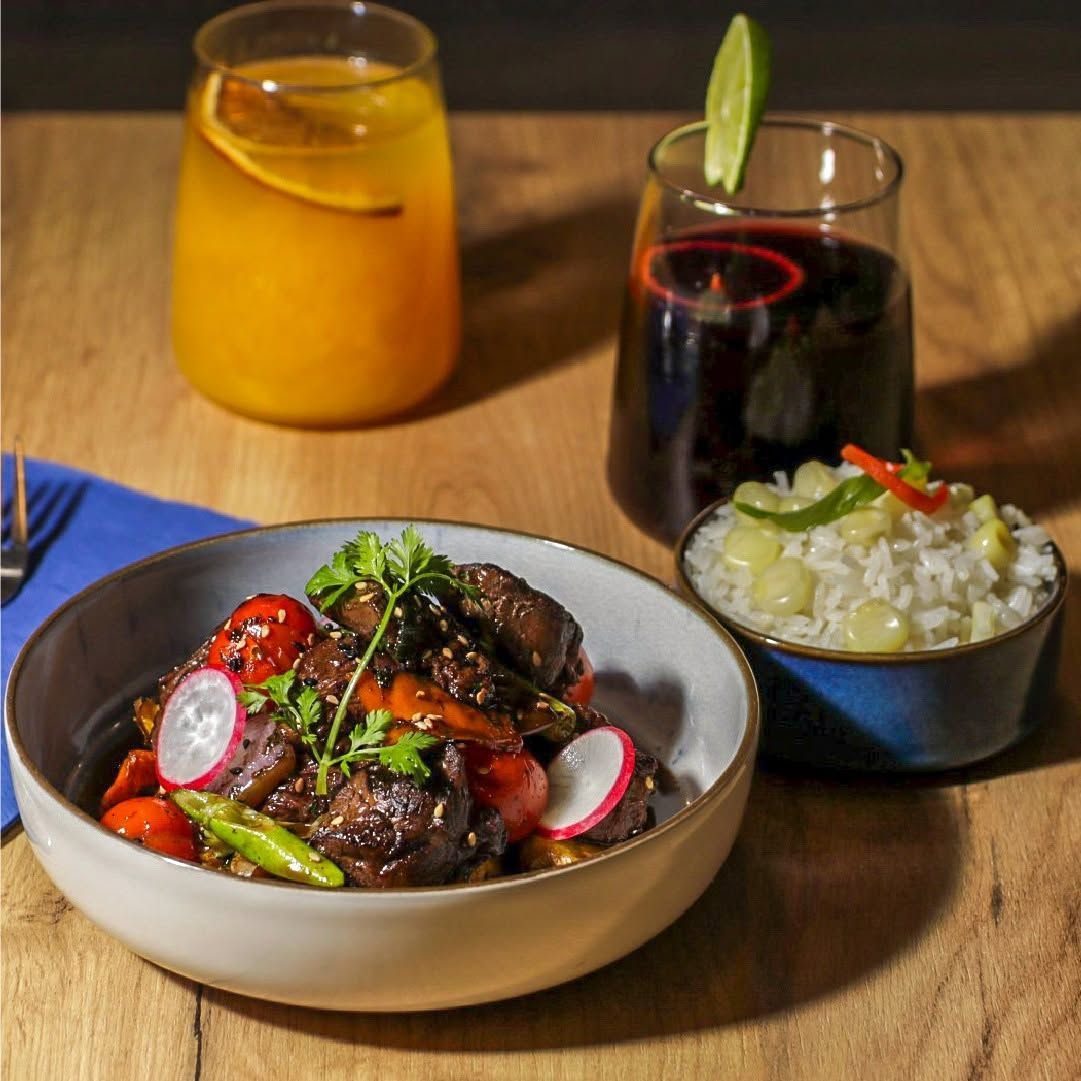 Plato de carne con salsa, rábanos y hierbas, acompañado de bebidas y una guarnición sobre una mesa de madera.