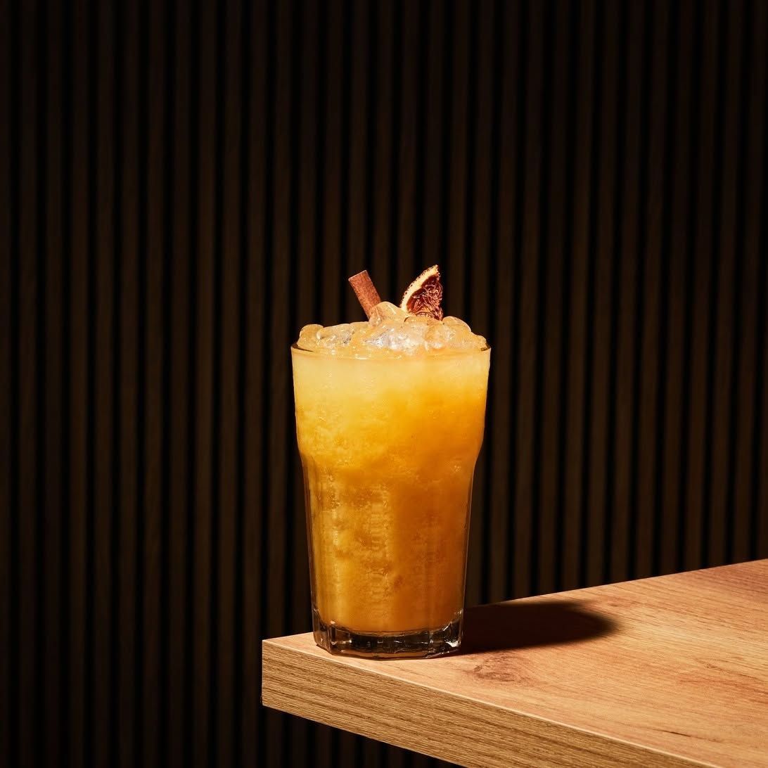 Cóctel alto de color naranja, con hielo y adornos, sobre una repisa de madera y con un fondo oscuro