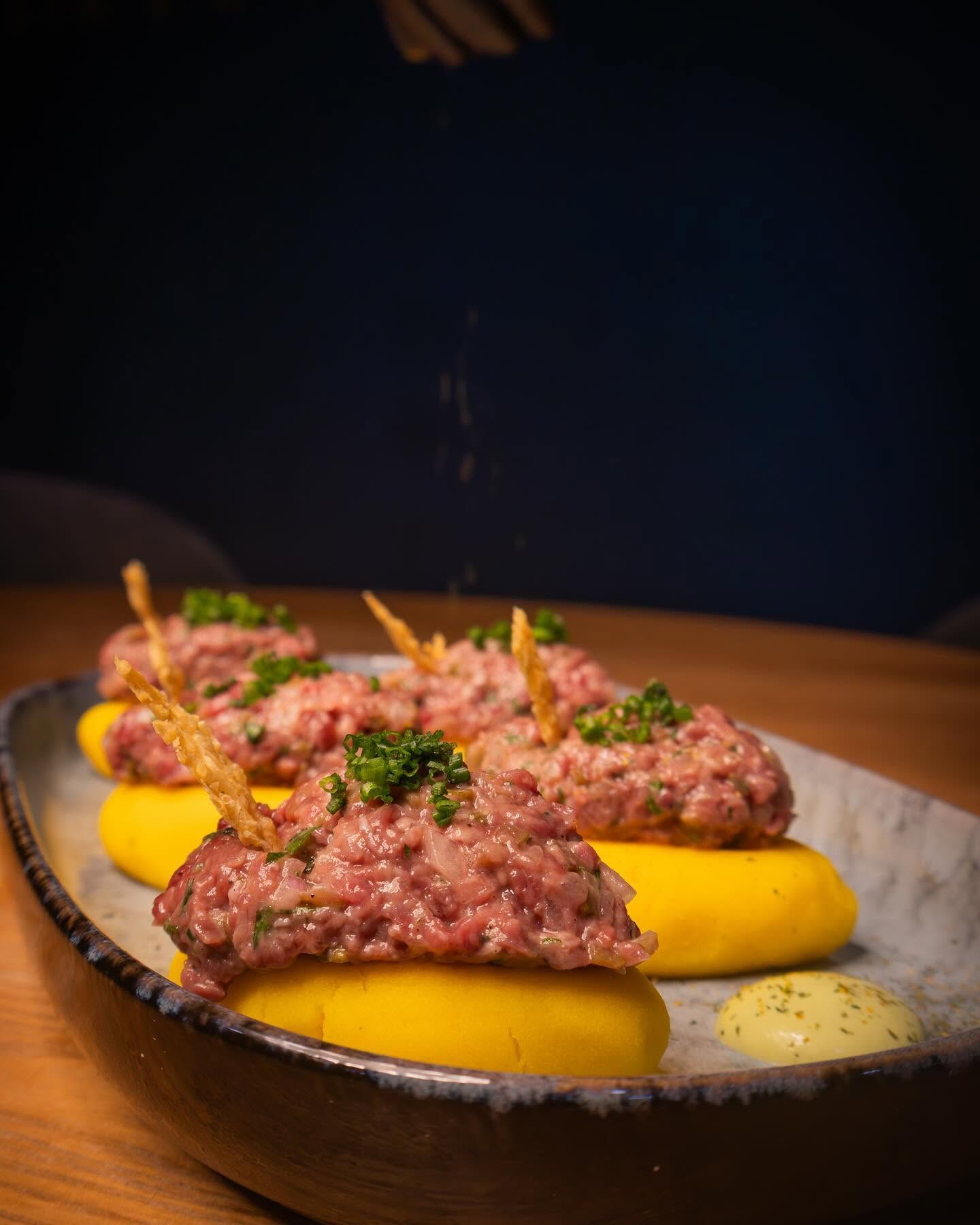 Plato gourmet emplatado con carne picada, puré amarillo, hierbas y salsa sobre una mesa oscura.