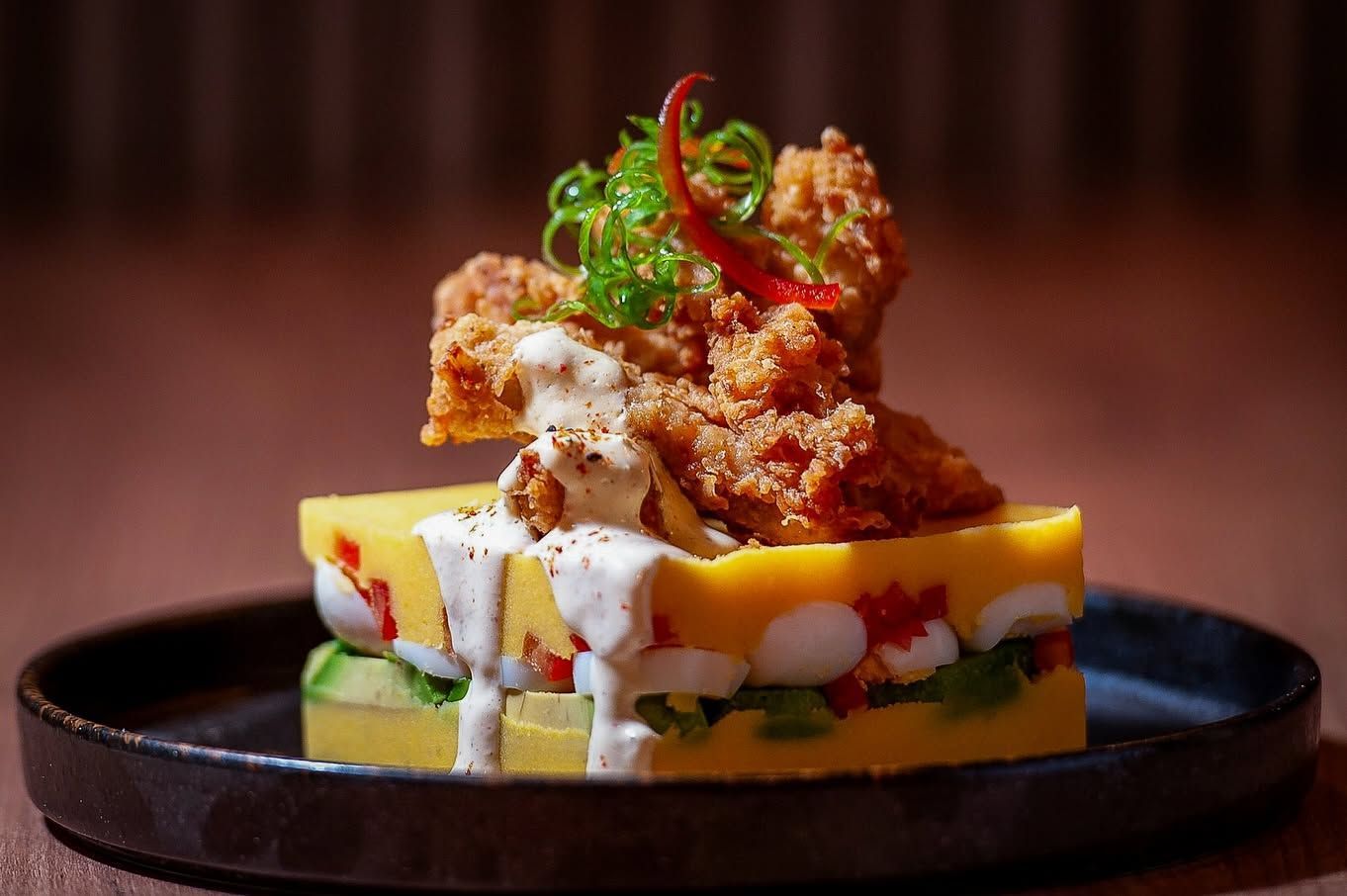 Pollo frito gourmet sobre capas de verduras amarillas y verdes en un plato negro, aderezado con hierbas frescas