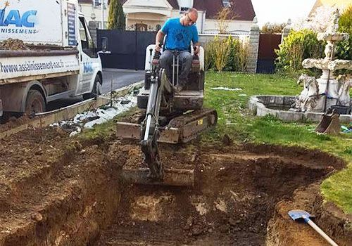 Une petite excavatrice creuse un grand trou dans un jardin résidentiel, un ouvrier la manœuvrant.