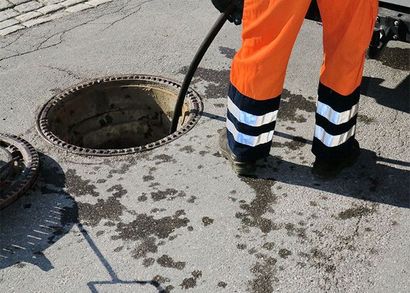 Un ouvrier en combinaison orange se tient près d'une plaque d'égout ouverte dans une rue mouillée.