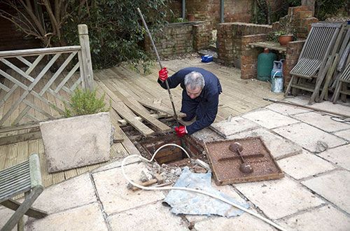 Une personne utilise une scie à perche au-dessus d'un caniveau ouvert dans une cour-terrasse, avec des outils et des pavés à proximité.