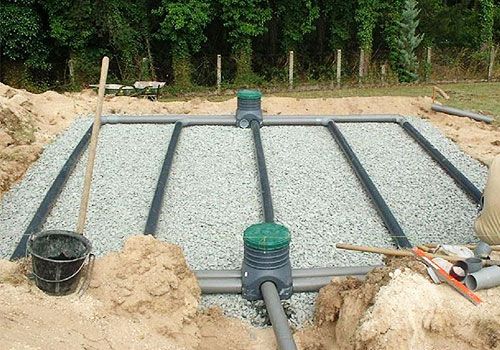 Lit de gravier pour champ d'épandage avec tuyaux de drainage noirs, un seau et des outils dans une zone de fouille.