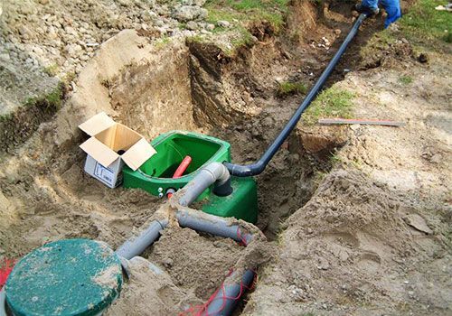 Un ouvrier installe un tuyau noir et un boîtier électrique vert dans une tranchée de terre, ses outils à proximité.