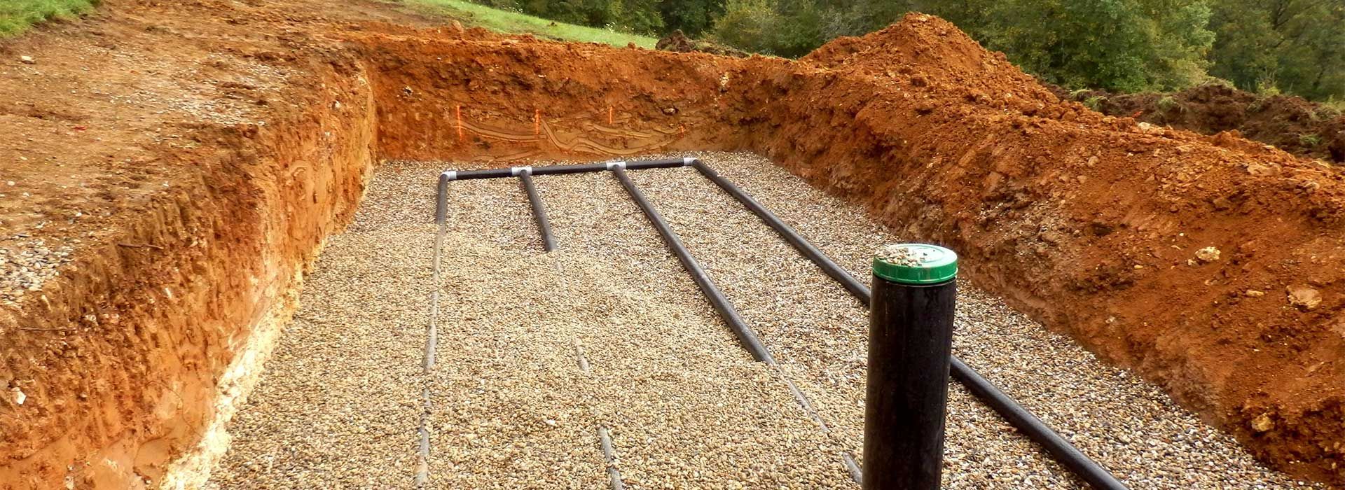 Tranchée de gravier avec des tuyaux noirs apparents et un poteau à capuchon vert dans un chantier de fouilles.