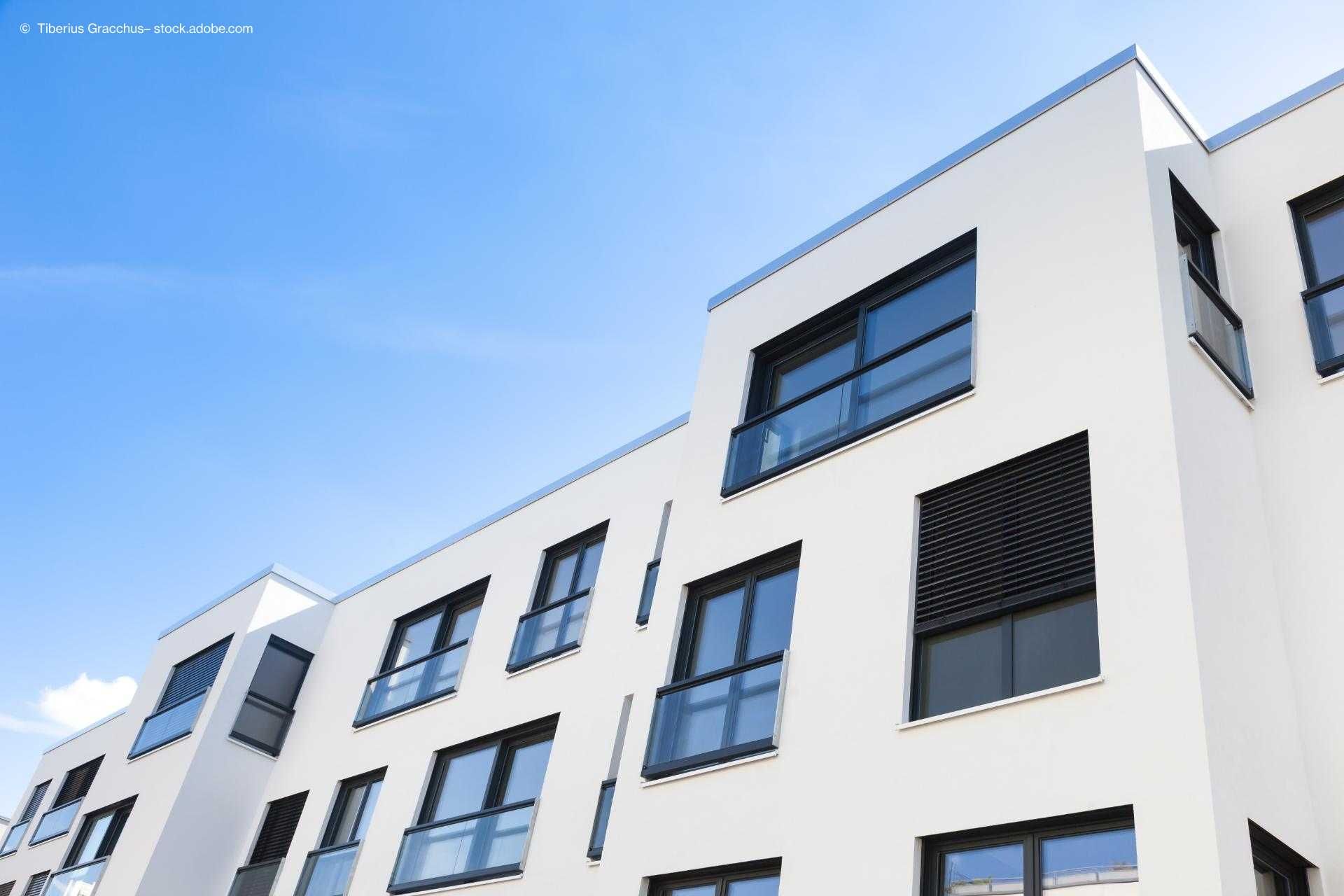 Modernes Wohngebäude mit weißer Fassade, großen Fenstern und Balkonen unter strahlend blauem Himmel.