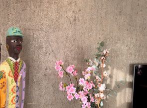 Bunte Figur neben einem dekorativen blühenden Zweig vor einer strukturierten Wand.