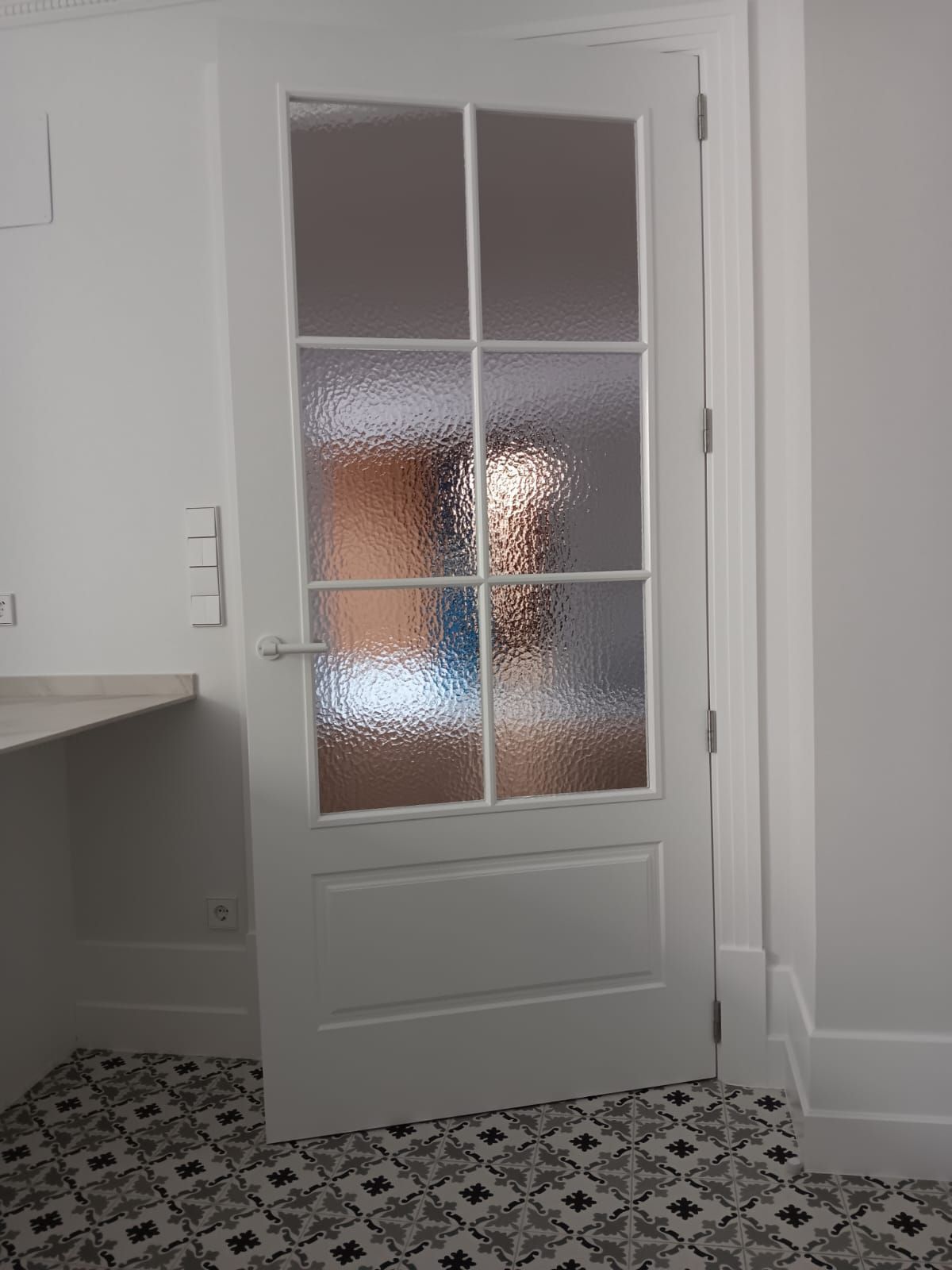 En el interior de una habitación, se observa una puerta interior blanca con paneles de vidrio esmerilado y un suelo de baldosas geométricas blancas y negras.