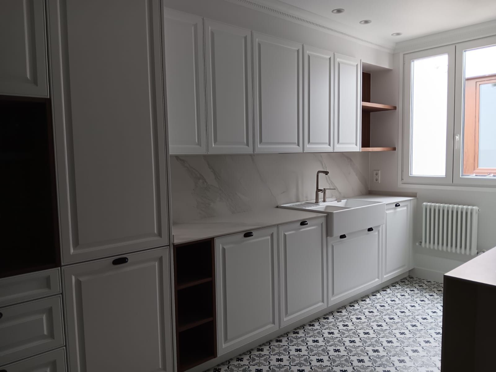 Cocina blanca y luminosa con armarios, fregadero, salpicadero de mármol y suelo de baldosas estampadas junto a una ventana.