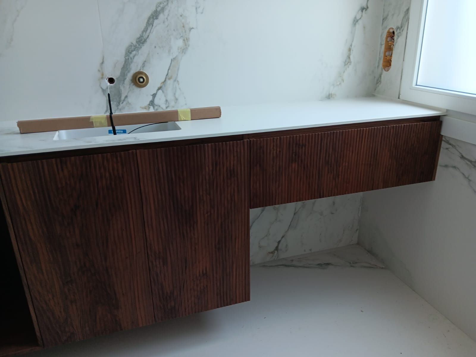 Tocador moderno de madera oscura con lavabo en un rincón de baño con azulejos de mármol blanco.