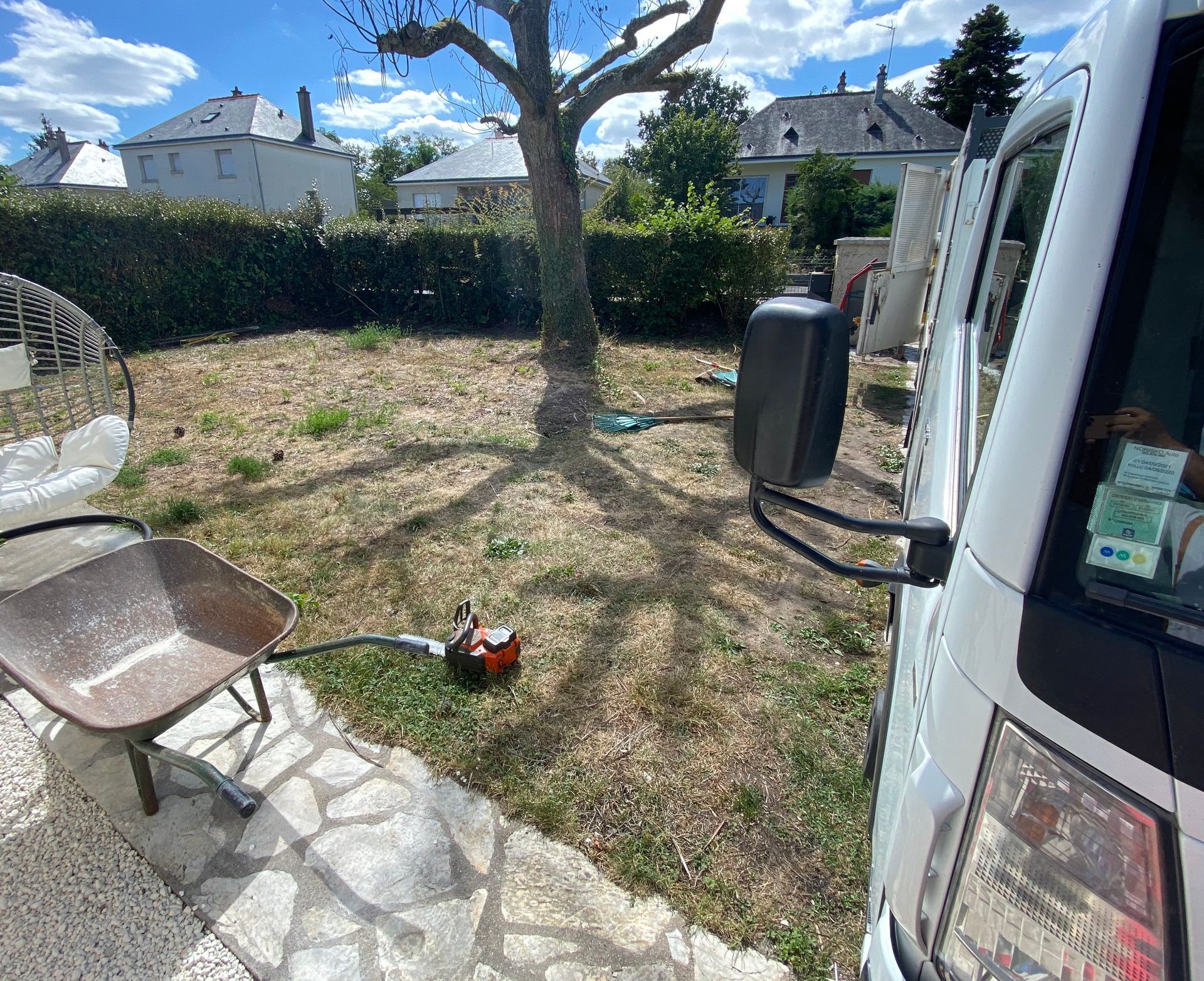 Vue d'un jardin à terrasser avec un arbre au milieu, le camion d'élagueur à droite, une brouette et un fauteuil à gauche