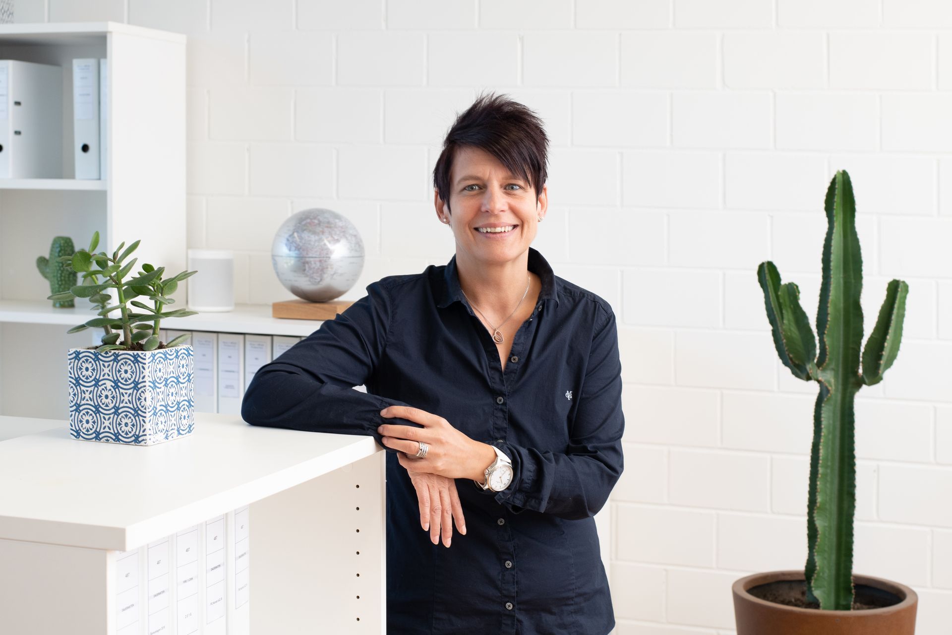 Frau mit kurzen Haaren, lächelnd, lehnt an einem weißen Schreibtisch in einem Büro mit einem Globus und einem Kaktus.