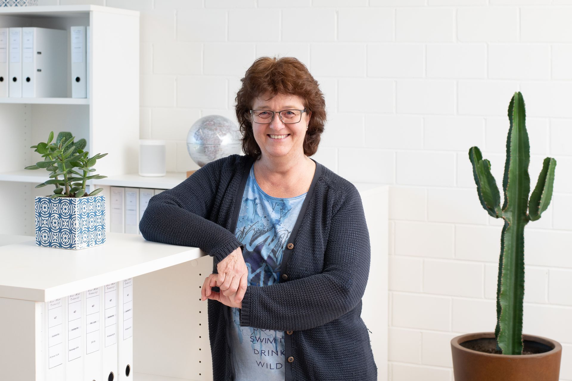 Frau mit Brille lächelt und lehnt an einem Schreibtisch in einem Büro mit einem Kaktus und einer Pflanze.