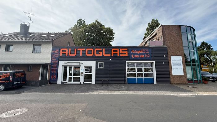 Autoglas-Geschäftsfassade mit großem orangefarbenem Schild, weißem Eingang und geparktem Fahrzeug neben einem Backsteingebäude.