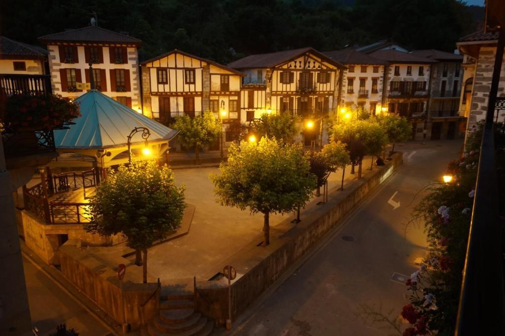 Vista nocturna de la plaza de un pueblo europeo con edificios iluminados, árboles y un mirador.