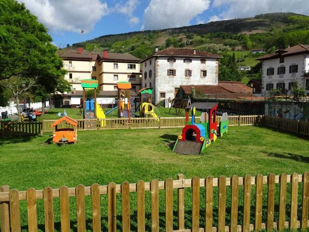 Parque infantil con equipamiento colorido, zona de césped, valla de madera y edificios al fondo.
