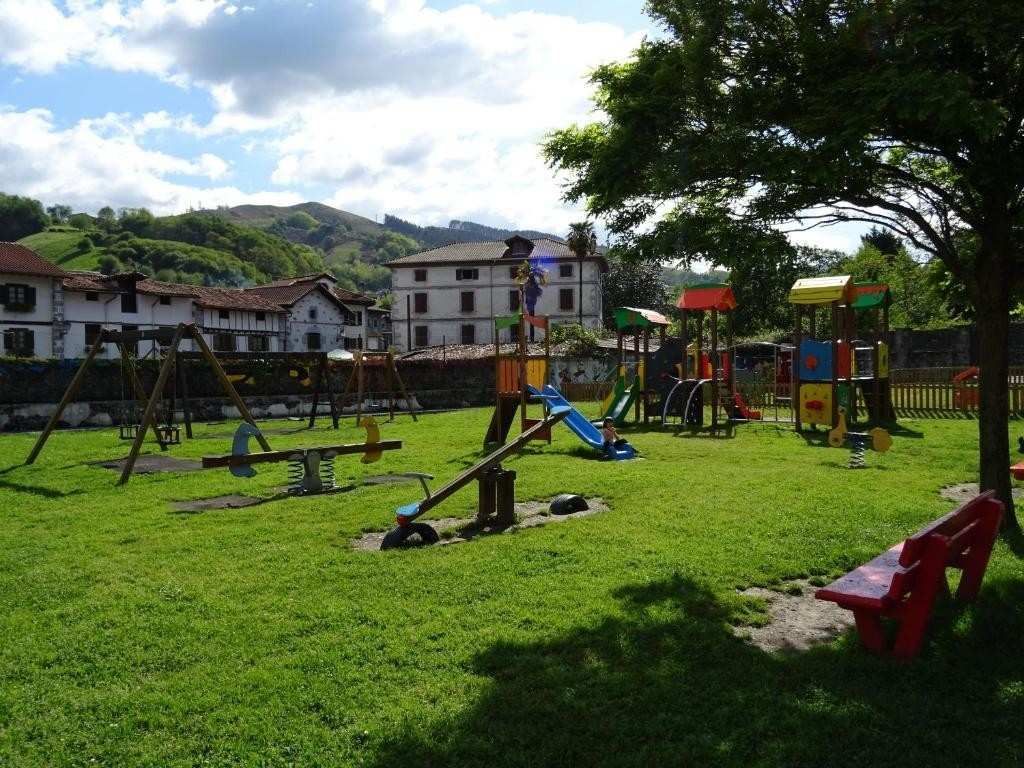 Parque infantil con césped verde, columpios, toboganes y estructuras de juego, con edificios y colinas al fondo.