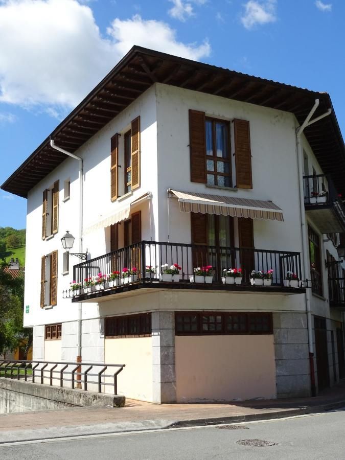 Edificio blanco de dos plantas con persianas marrones y balcón. Base de piedra, toldo y flores.