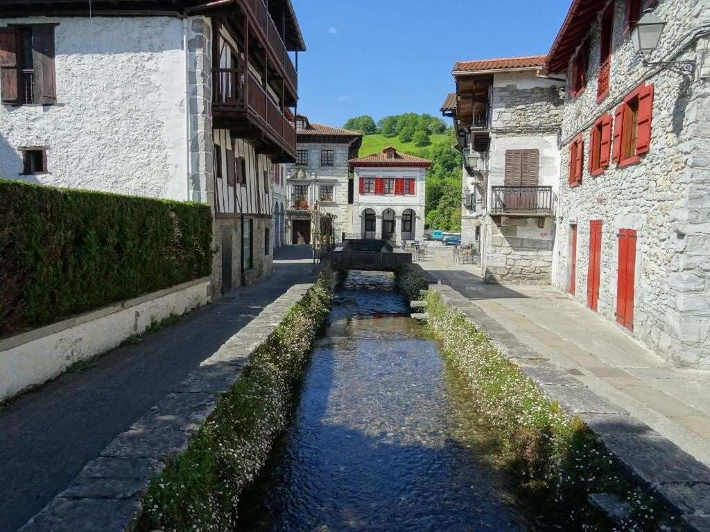 Calle con edificios de piedra, un arroyo y contraventanas rojas, con un fondo de colinas verdes.