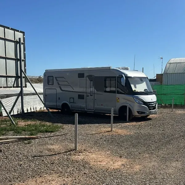 Una autocaravana plateada aparcada sobre grava junto a un edificio bajo un cielo azul despejado.