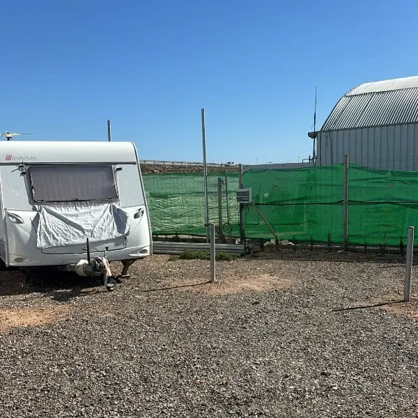 Caravana blanca junto a una valla de malla verde en un terreno de grava bajo un cielo azul despejado.