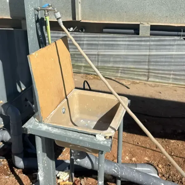 Fregadero exterior con tapa de madera, estructura metálica y mangueras junto a la pared de un edificio.