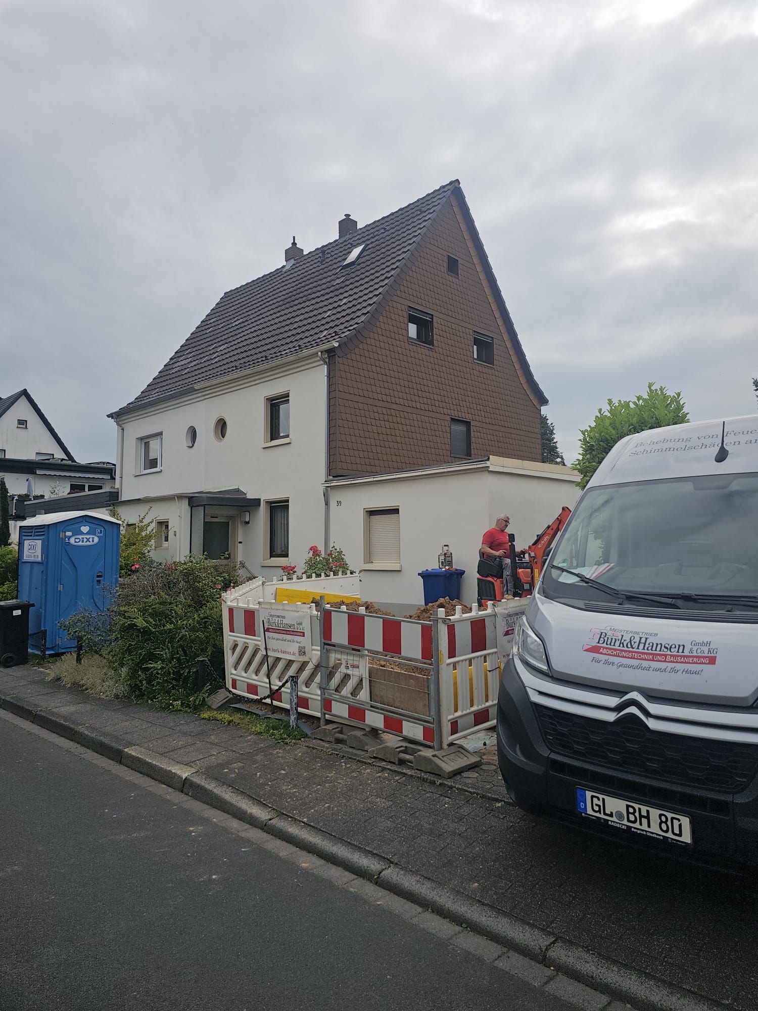 Straßenansicht eines im Bau befindlichen Hauses mit Absperrungen, einer blauen Mülltonne und einem daneben geparkten weißen Lieferwagen.