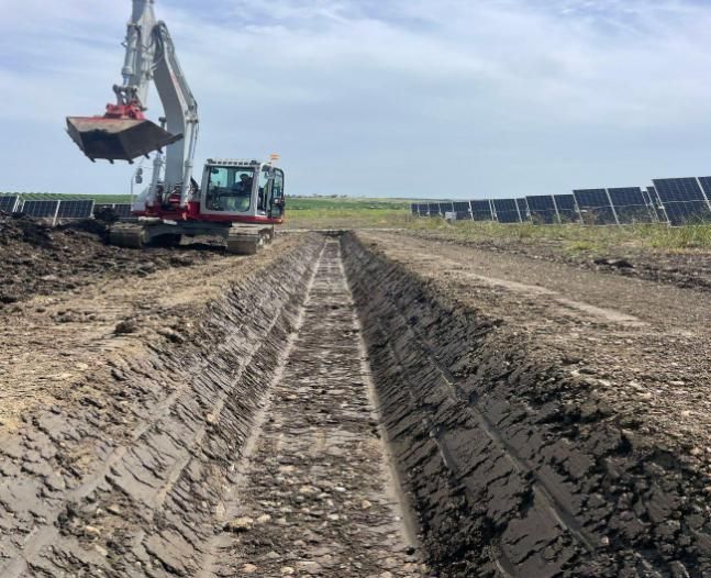 Excavadora cavando zanja cerca de paneles solares en el campo bajo un cielo nublado.