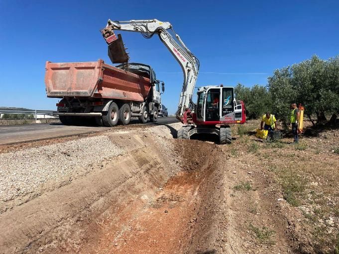 Una excavadora carga un camión volquete con tierra al borde de la carretera. Trabajadores con chalecos amarillos cerca.