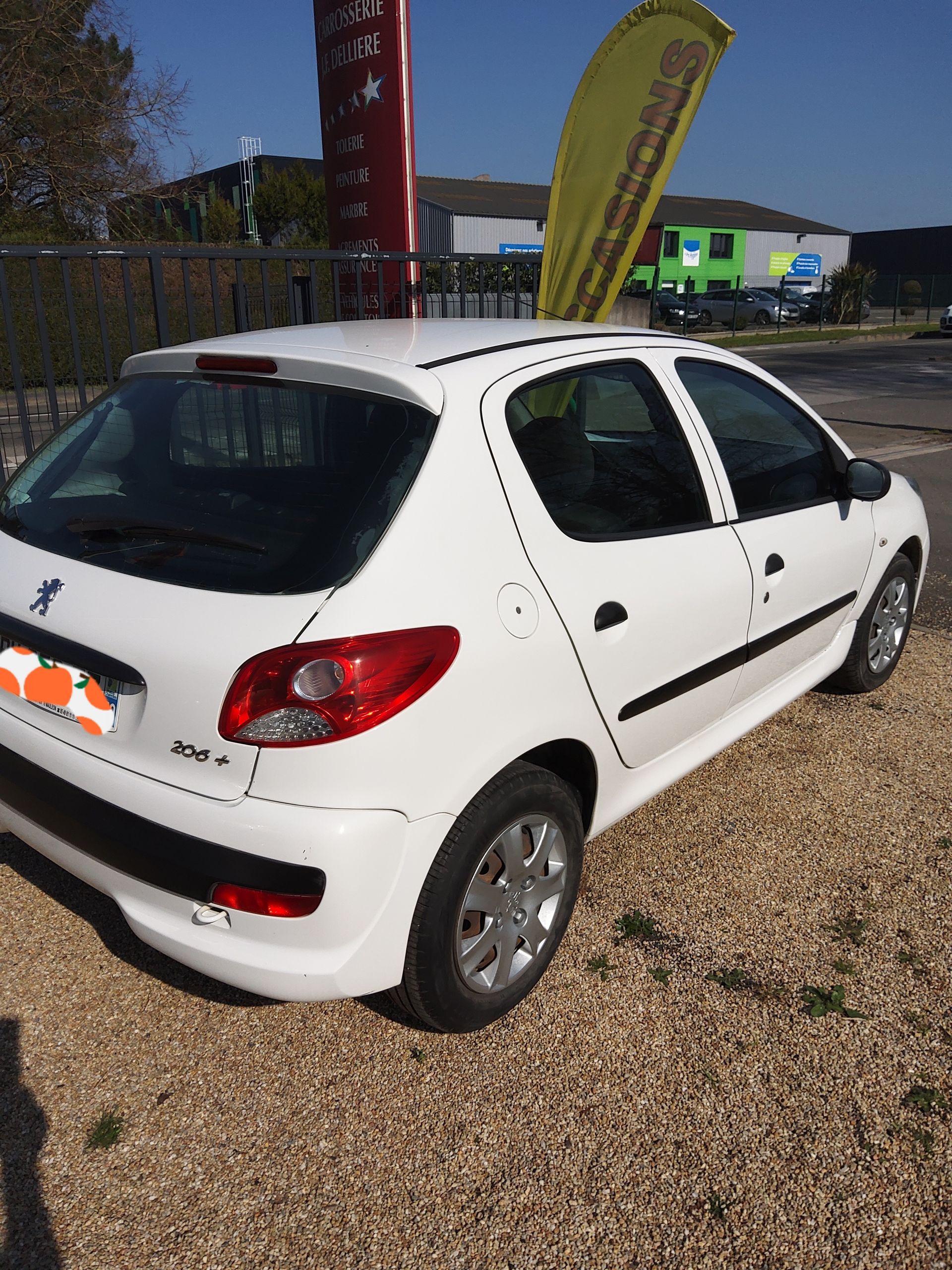 Une Peugeot 206+ blanche garée sur un parking gravillonné à côté d'un drapeau publicitaire jaune