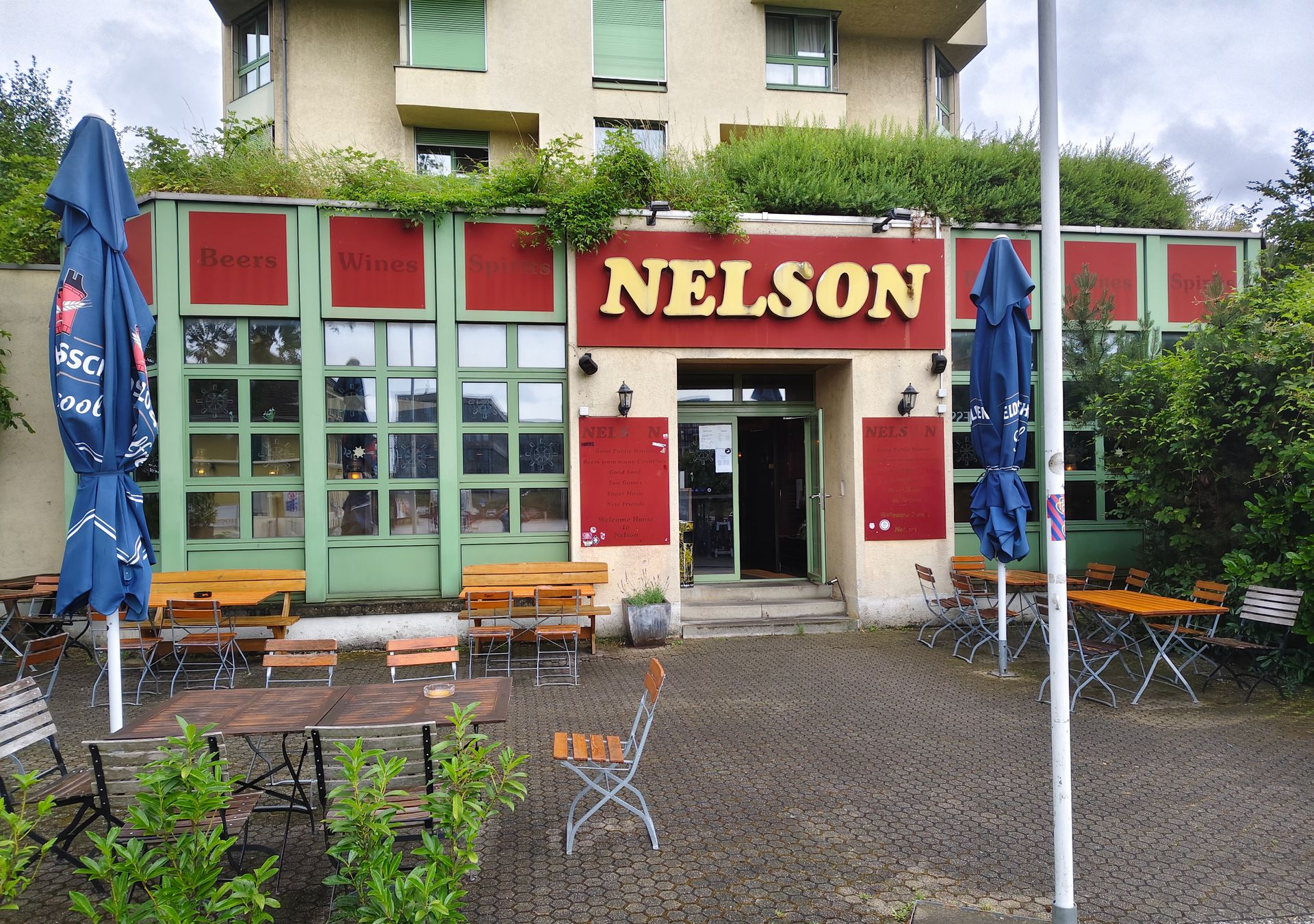 Schaufenster mit einem roten „NELSON“-Schild, Fahnen und einem darüberliegenden Gebäude, von der Strasse aus gesehen.