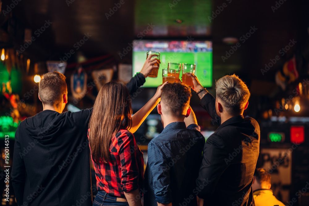 Freunde jubeln und erheben ihre Getränke in einer Sportsbar, während sie ein Fussballspiel auf einem Fernsehbildschirm verfolgen.