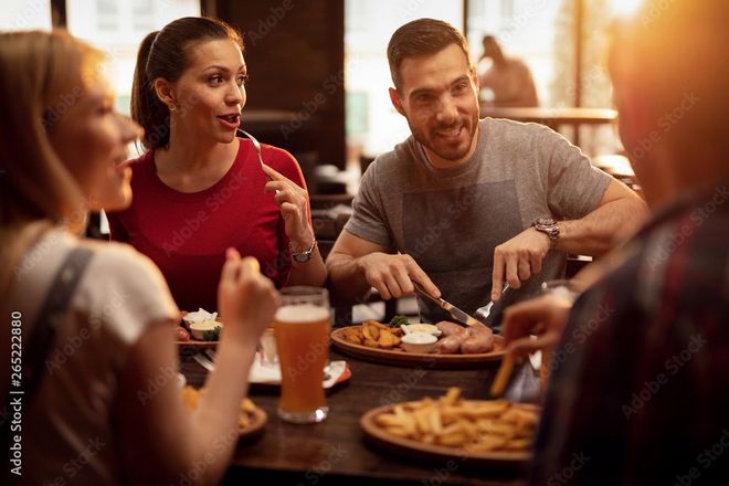 Freunde unterhalten sich beim Abendessen in einem gemütlich beleuchteten Restaurant; Getränke, Pommes frites und eine gemeinsam zubereitete Speise stehen auf dem Tisch.