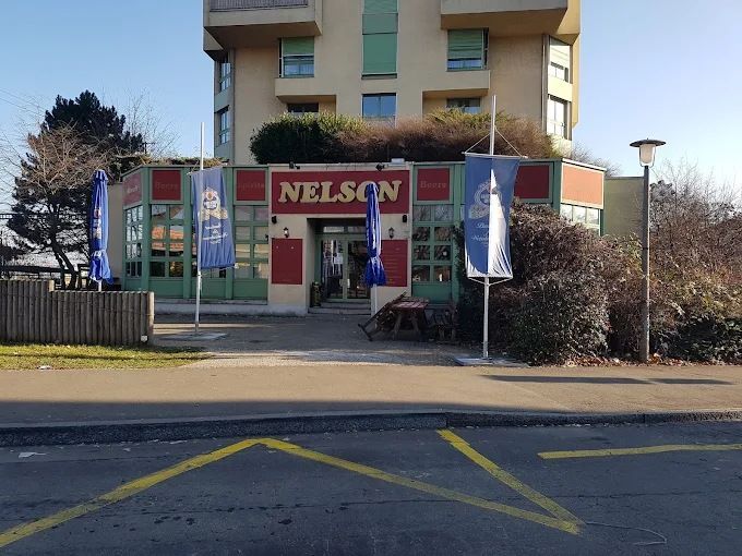 Schaufenster mit einem roten „NELSON“-Schild, Fahnen und einem darüberliegenden Gebäude, von der Strasse aus gesehen.