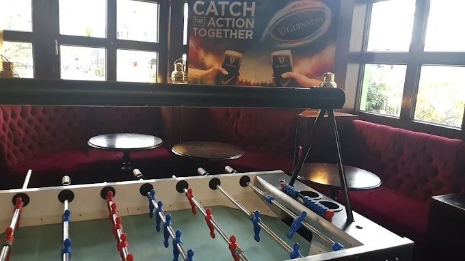 Ein Tischkicker in einem Pub mit roten Sitznischen, runden Tischen und einem „Catch Action Together“-Poster an der Wand