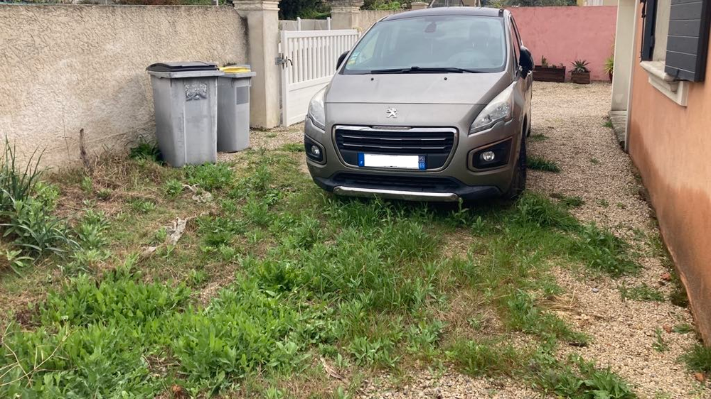 Un SUV gris garé dans une allée de gravier envahie par les hautes herbes, à côté de poubelles et d'une maison.