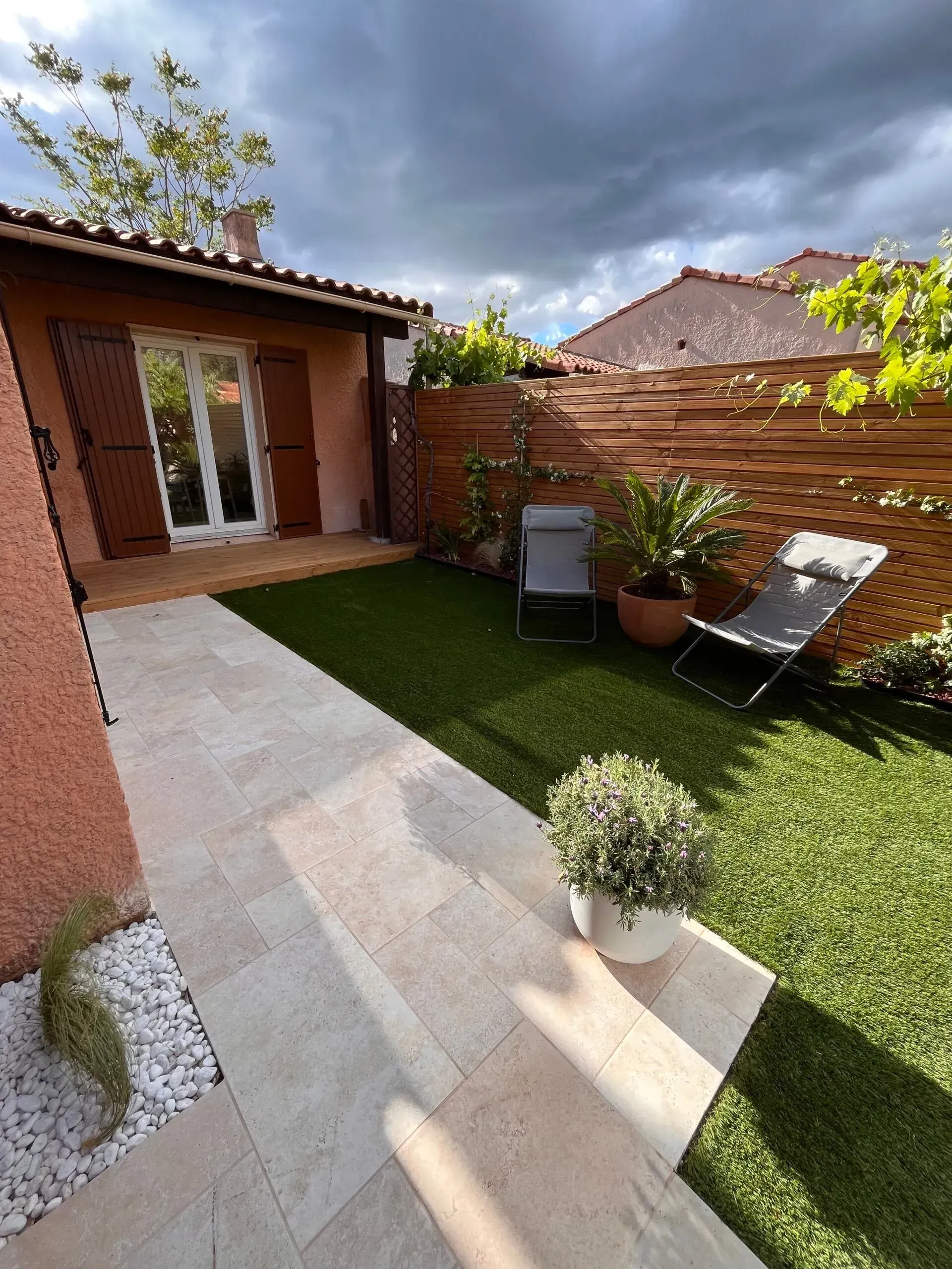 Petit jardin ensoleillé avec un chemin, une pelouse verte, une clôture en bois et une chaise longue.