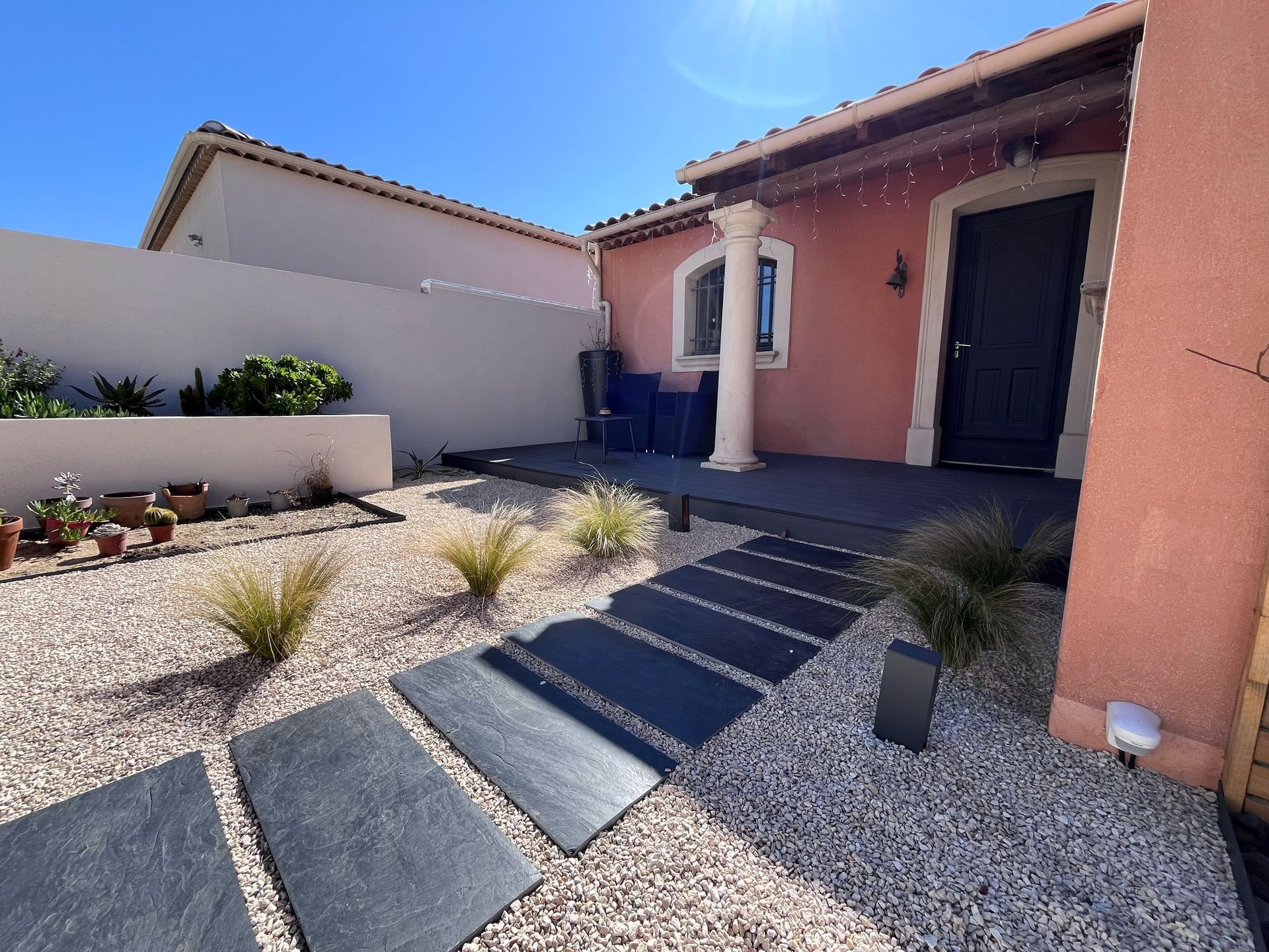 Allée de dalles sombres traversant une cour de gravier menant à l'entrée d'une maison. Journée ensoleillée.