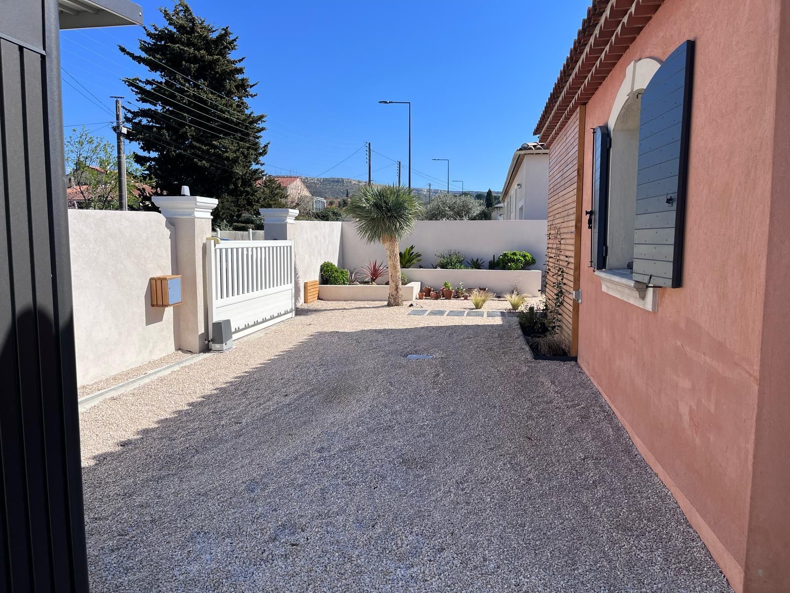 Une allée de gravier mène à un portail blanc et à une maison rose aux volets noirs. Journée ensoleillée, ciel bleu.