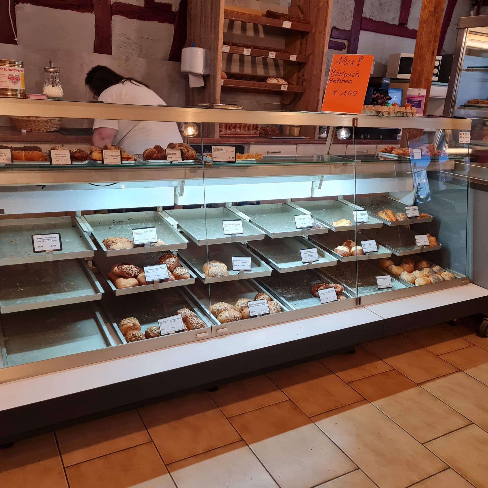 Auslage einer Bäckerei mit Gebäck. Eine Person arbeitet hinter der Theke.