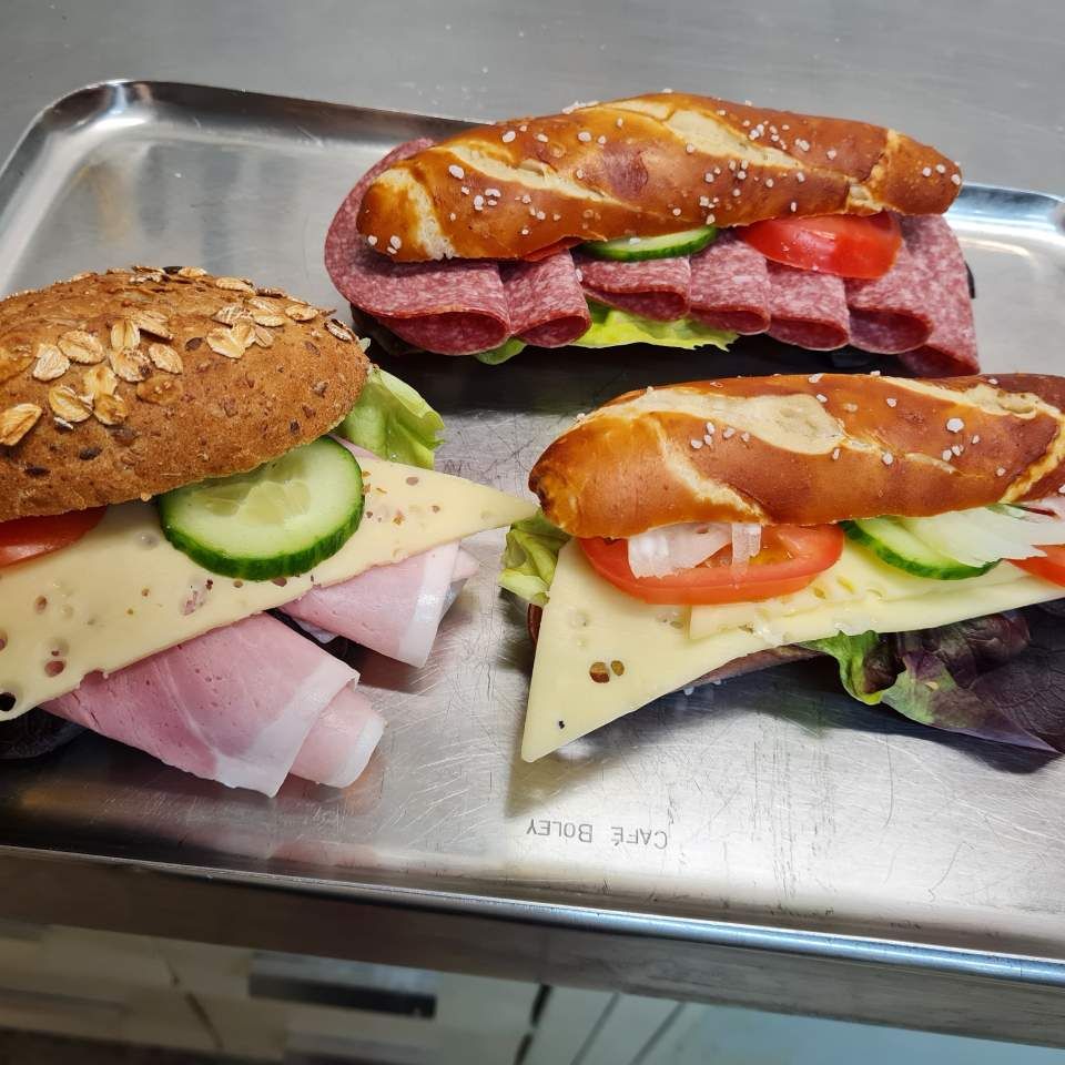 Drei Sandwiches auf einem Metalltablett: Schinken und Käse auf einem Sesambrötchen, Salami auf einem Brezelbrötchen und Käse, Tomate und Gurke auf einem Brezelbrötchen.