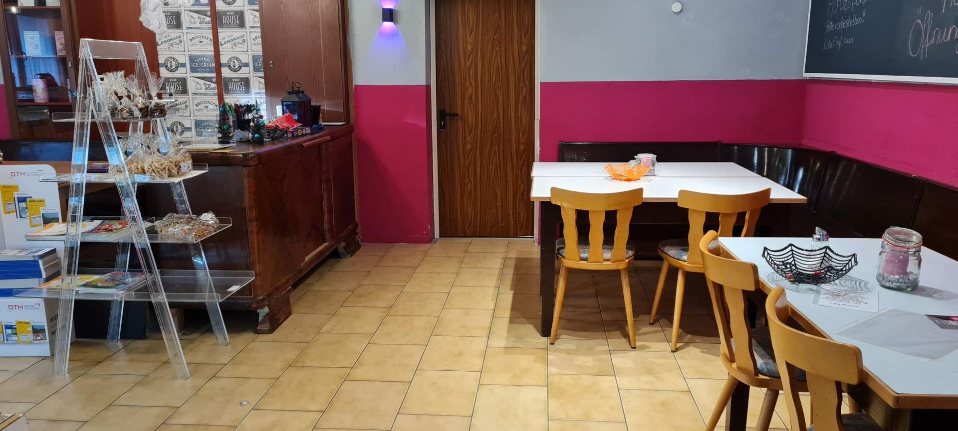 Innenansicht eines Restaurants mit Holztischen und -stühlen. Rosa Wände, eine Holzbar und Fliesenboden sind zu sehen.