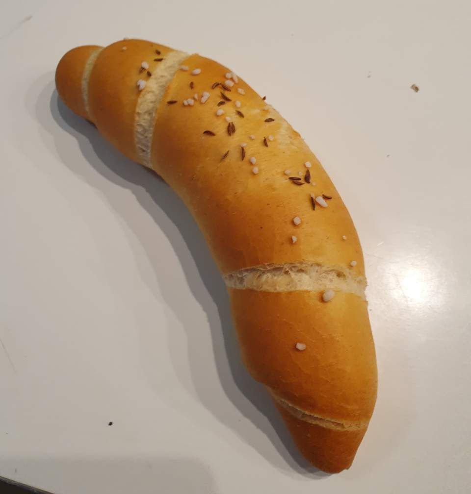 Ein gebogenes, goldbraunes Brötchen, bestreut mit Körnern und Salz, auf weißem Untergrund.