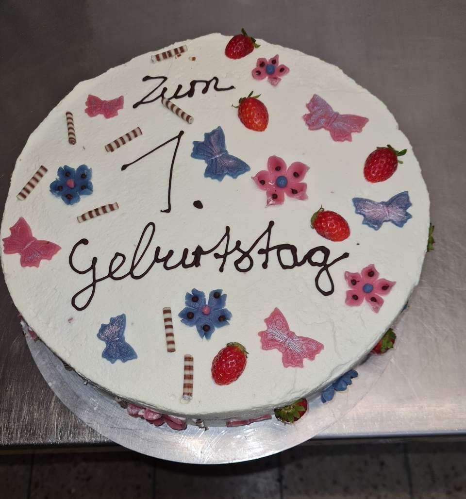 Kuchen mit weißem Zuckerguss, verziert mit Schmetterlingen, Blumen, Erdbeeren und dem Schriftzug „Zum 1. Geburtstag“ in dunkler Schrift.