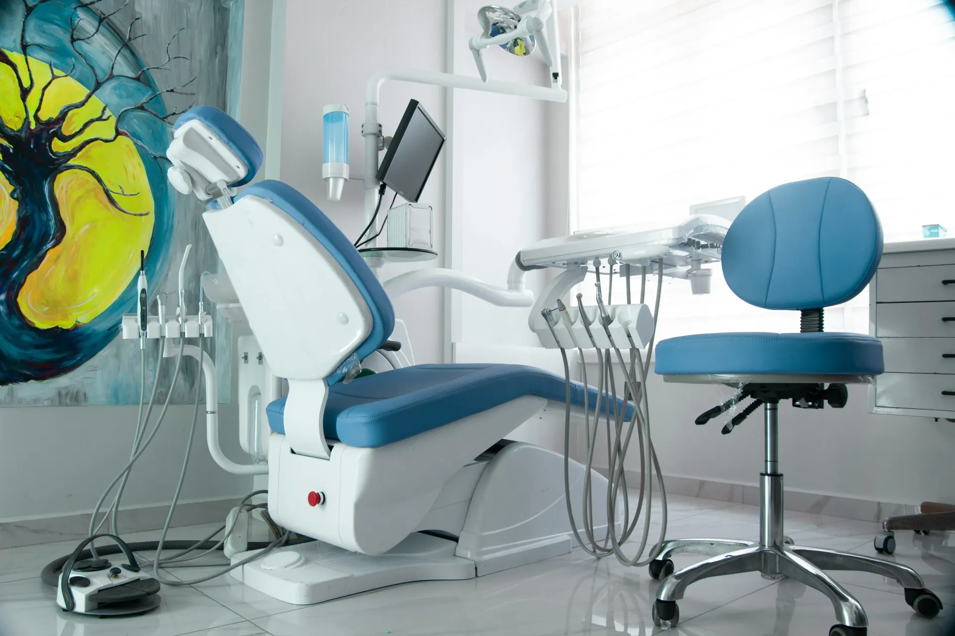 Sala de examen dental con una silla azul y blanca, herramientas y un taburete de dentista.