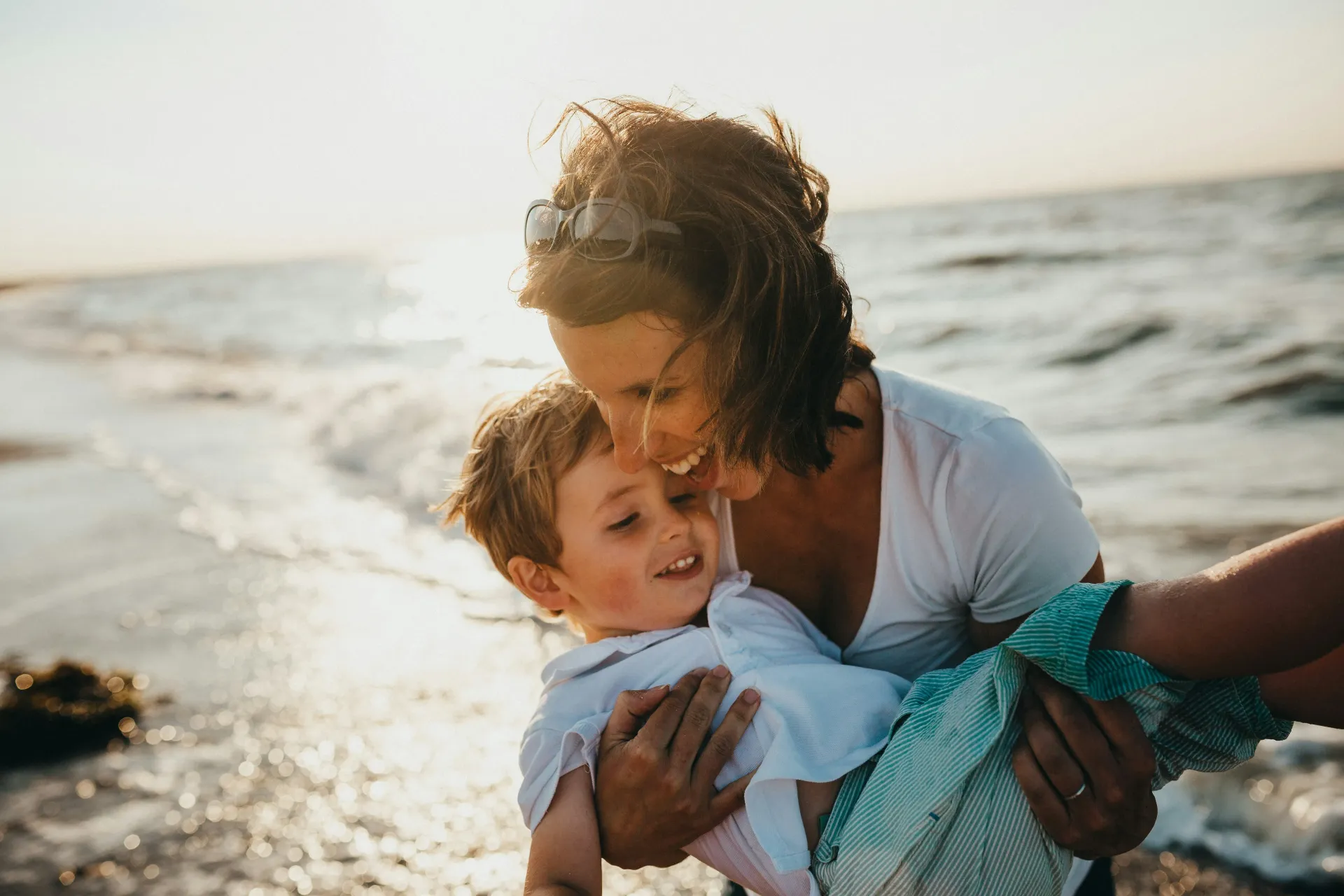 Una mujer abraza y besa a un niño en la playa. Ambos sonríen, iluminados por el sol.