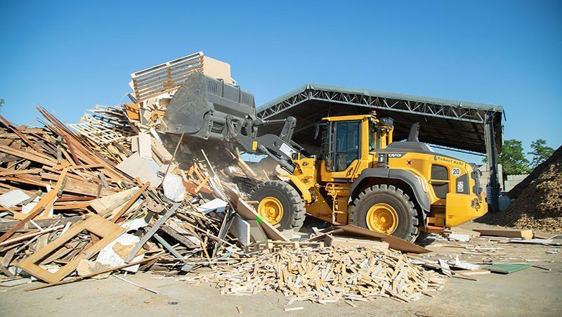 Gelber Lader hebt Holzabfälle in einer Recyclinganlage unter blauem Himmel.